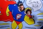 Ojciec i syn, których jedynie głowy widać na tle komiksowej sylwetki supermena, trzymającego w ręku małe dziecko. W komiksowym dymku są słowa wypowiadane przez dziecko: Super tata!