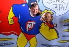 Ojciec i córka, których głowy widać na tle komiksowej sylwetki supermena trzymającego w ręku małe dziecko