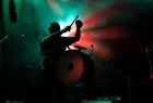 Perkusista trzyma uniesione pałeczki nad bębnem. Za nim widać zielone i czerwone światła. 