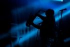 Ciemna scena podświetlona niebieskimi snopami światła, w tle widać sylwetkę mężczyzny grającego na saksofonie. 