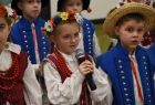 Dziewczynka ubrana w ludowy strój z kolorowym wiankiem na głowie trzyma mikrofon, obok chłopcy, ubrani w niebieskie marynarki i słomiane kapelusze. Występ przedszkolaków w nowym przedszkolu w Sękowej. 