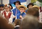 Chłopczyk ubrany w niebieską marynarkę i słomiany kapelusz, w ręku trzyma mikrofon, w tle kilkoro dzieci w strojach ludowych. Dzieci występują podczas otwarcia nowego przedszkola w Sękowej. 