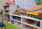Sala nowego przedszkola, na pierwszym planie widać biało - zielono - żółtą szafkę, z kolorowymi zabawkami dziecięcymi. Szafa stoi przy ścianę pokrytej kolorowym rysunkiem z motywami roślinnymi. 