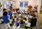 Grupa przedszkolaków siedzących w koło na kolorowym dywanie w sali przedszkolnej. Dzieci ubrane galowo część w strojach ludowych. Na ścianach znajdują się dziecięce rysunki i szafy z kolorowymi zabawkami i pomocami naukowymi. 