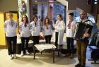 7 osób z zespół Ruczaj, śpiewa Kolendy łemkowskie. Zespół to 5 kobiet i 2 mężczyzn, mają na sobie białe koszule z tradycyjnym łemkowskim haftem. Na pierwszym planie widać akordeonistę. Zespół stoi w holu domu wczasowego w Wapiennem gmina Sękowa.