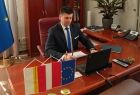 Wicemarszałek Łukasz Smółka siedzi przy stole w gabinecie i patrzy w monitor komputera. Na pierwszym planie widoczne flagi Małopolski, Polski i Unii Europejskiej.