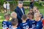 Marszałek Witold Kozłowski stoi w otoczeniu kilkunastu dzieci. Dzieci stoją tyłem do zdjęcia, mają na sobie niebieskie koszulki piłkarskie z numerami. Marszałek wita się z jednym z chłopców. Wszyscy stoją na zielonej murawie boiska, w tle widać rodziców.