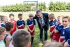 Marszałek Witold Kozłowski stoi w kręgu z młodszymi piłkarzami. Dzieci maja na sobie niebiesko czerwone stroje. Marszałek pochyla się do chłopców w tle widać zieloną murawę boiska, białą bramkę piłkarską i zielone drzewa. 