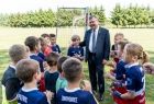 Marszałek Witold Kozłowski stoi na zielonej murawie boiska za nim znajduję się biała bramka piłkarska. Do około marszałka ustawiła się grupa uśmiechniętych dzieci, w tle widać białą bramkę piłkarską i zielone drzewa. Dzieci ubrane są w stroje piłkarskie.