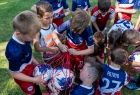 Grupa dzieci w piłkarskich strojach, pochyla się nad workiem wypełnionym piłkami. Część chłopców stoi część siedzi na murawie, dzieci są uśmiechnięte. Chłopcy ubrani są w niebiesko czerwone stroje piłkarskie. 