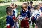 Grupa dzieci w piłkarskich niebiesko czerwonych strojach, dwóch chłopów otwiera szary worek z prezentami od marszałka Witolda Kozłowskiego. Chłopcy uśmiechają się. Cała grupa stoi na zielonym piłkarskim boisku. 