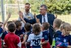 Marszałek Witold Kozłowski, przybija piątki z młodymi piłkarzami. Dzieci ubrane są w niebiesko czerwone stroje piłkarskie. Z prawej strony za marszałkiem stoi trener za nim widać białą bramkę piłkarską, zieloną murawę i zielone drzewa. 