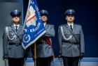 Trzech policjantów w mundurach stoi na baczność. Stojący w środku policjant trzyma sztandar.