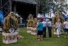 Widok na 3 wieńce dożynkowe ustawione na kwadratowych podstawach,. Wieńce ustawione są przy scenie na trawniku. W śród nich widać spacerujących ludzi, w tle widać drzewa i niebieskie niebo.