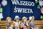 Dzieci biorą udział w występie artystycznym i siedzą na ławce. W tle widoczny napis: W dniu Waszego święta.