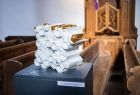 Widok na prace Stanisława Brach. Jest to biała rzeźba w kształcie klastra miodu ze złotymi elementami. Praca umieszczona jest na podeście we wnętrzu kościoła