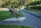 Widok na pomnik Jana Kiepury, ustawiony w śród miejskiej zieleni przy wąskiej alejce. Pomnik ma kształt ławeczki na której siedzi postać wybitnego artysty. W tle widać zielone drzewa. 