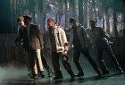 Widok na tańczących artystów. Mężczyźni pochylają się w prawą stronę. Scena jest delikatnie podświetlona, do minują na niej kolory szarości. 