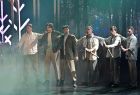 Widok na tańczących artystów. Mężczyźni pochylają się w prawą stronę. Scena jest delikatnie podświetlona, do minują na niej kolory szarości. Scena spowija delikatna mgła.