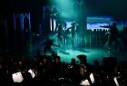 Widok na scenę na której wsypują artyści, scena pokryta jest bladoniebieskim światłem i sztuczną mgłą. Przy scenie widać muzyków. 
