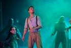Artyści w trakcie występu na scenie Opery Krakowskiej. Na pierwszym planie widać młodego mężczyznę ubranego w wiejski strój. Zanim w idać pozostałych artystów scena podświetlona jest na blado niebieski kolor. 