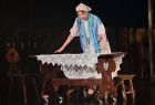 Aktorka stoi na scenie, ubrana jest w strój gospodyni, ręce trzyma na drewnianym stole na którym rozłożono biały haftowany obrus. 