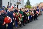 Uczestnicy uroczystości niepodległościowych w Brzesku stoją wzdłuż ulicy, trzymając kwiaty i wieńce