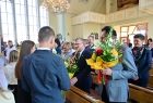 Grupa uczniów w odświętnych stroją stoi wewnątrz kościoła, młody mężczyzna trzyma w ręku bukiet kwiatów. W tle widać fragment organów kościelnych i wysokie witrażowe okna. 