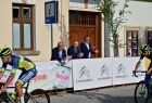 Widok na kolarza jadącego na rowerze, za nim w tle widać Marszałka Witolda Kozłowskiego i burmistrza starego Sącza Jaka Lelka. W tle za nimi widać ściany kamienic, przed nimi banery promujące partnerów wyścigu.