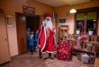 Święty Mikołaj wraz z grupka dzieci wchodzi do pomieszczenia wypełnionego prezentami. 