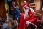 Mikołaj stoi w otoczeniu grupy dzieci. W tle Marszałek Małopolski przekazuję dzieciom prezenty. 