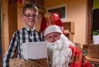 Chłopczyk w okularach i Święty Mikołaj pozują wspólnie do zdjęcia, chłopiec trzyma w ręku prezent. 