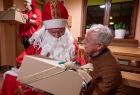 Starszy mężczyzna odbiera z rąk Świętego Mikołaja świąteczny prezent. 