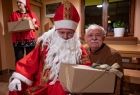 Starszy mężczyzna pozuje do wspólnego zdjęcia ze Świętym Mikołajem, mężczyzna trzyma w ręku świąteczny prezent. 
