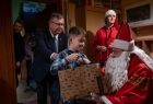 Chłopczyk w koszuli w kratę odbiera z rąk Świętego Mikołaja świąteczny prezent. Obok nich stoi Marszałek Witold Kozłowski. 