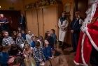 Grupa dzieci siedzi na podłodze przed Świętym Mikołajem, dzieci oglądają otrzymane od niego prezenty świąteczne. 