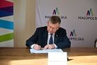 Marszałek Małopolski siedzi przy stole i podpisuję dokument dot. utworzenia ZAZ w Łukowicy. W tle widać logo małopolski umieszczone na białej ściance oraz fragment kolorowego loga.