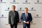 Marszałek Małopolski Witold Kozłowski i lokalny samorządowiec. Mężczyźni stoją na tle białej ścianki z napisami małopolska i kolorowym logiem w kształcie litery M.