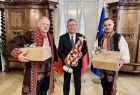 marszałek Witold Kozłowski w towarzystwie dwóch mężczyzn w strojach regionalnych, stoją na tle flag trzymając w ręce prezenty.