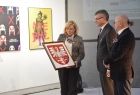 białe pomieszczenie w którego centralnym miejscu stoi radna Marta Mordarska, w ręku trzyma godło małopolski, obok niej stoi dyrektor galerii i burmistrz Jacek Lelek. W tle na białej ścianie zawieszone są dwa obrazy.