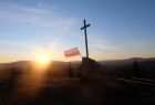 Flaga RP i krzyż na tle nieba i wschodzącego słońca