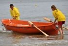 Ratownicy wodni w żółtych koszulkach wpychają pomarańczową łódź z wiosłami do wody.