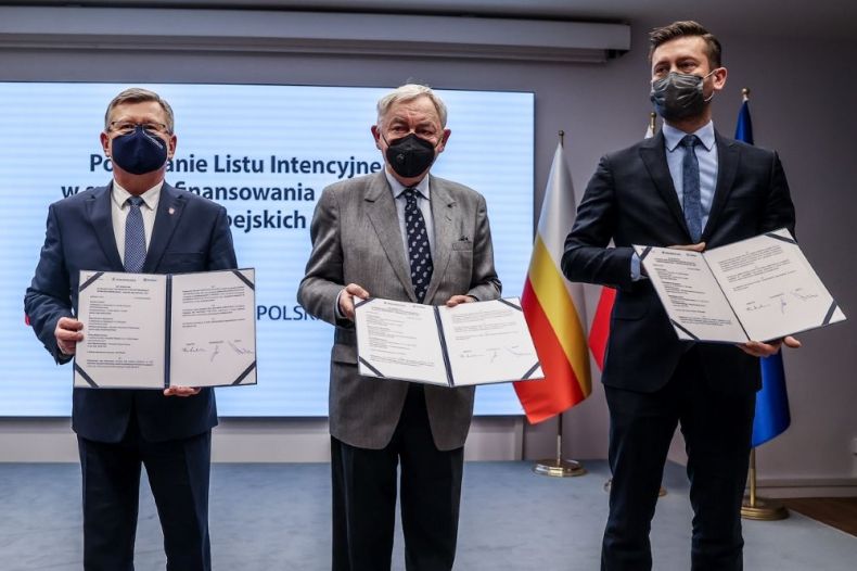 Marszałek Witold Kozłowski, prezydent Jacek Majchrowski i minister Kamil Bortniczuk stoją trzymając w rękach podpisane dokumenty. W tle widoczny ekran.