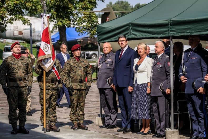 Iwona Gibas z zarządu województwa stoi na trybunie honorowej w muzeum lotnictwa. Obok inni ludzie, w tle widoczne samoloty.