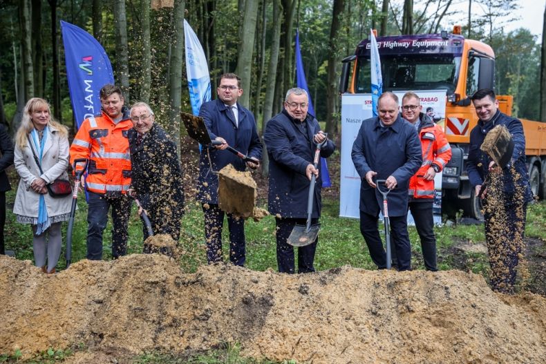 Uczestnicy wydarzenia wbijają łopaty w ziemię. Z tyłu widoczne flagi z napisem Małopolska i Fundusze Europejskie.