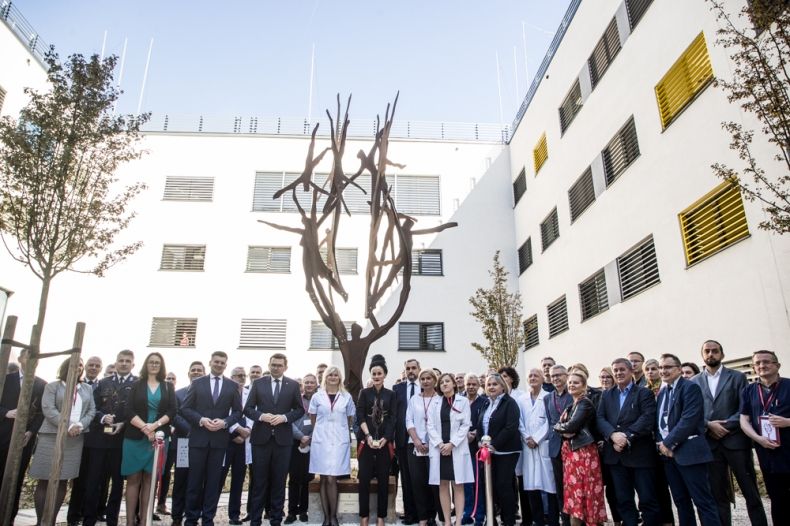 Uczestnicy wydarzenia stoją przed pomnikiem w kształcie drzewa na dziedzińcu szpitala.