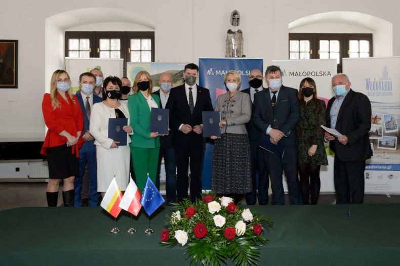 Uczestnicy wydarzenia stoją wewnątrz zabytkowego wnętrza. Na pierwszym planie widoczne kwiaty i flagi.