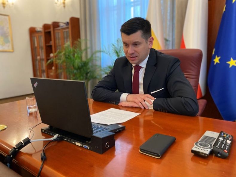 Wicemarszałek Łukasz Smółka siedzi przy biurku w gabinecie. Na pierwszym planie i w tle widoczne flagi.