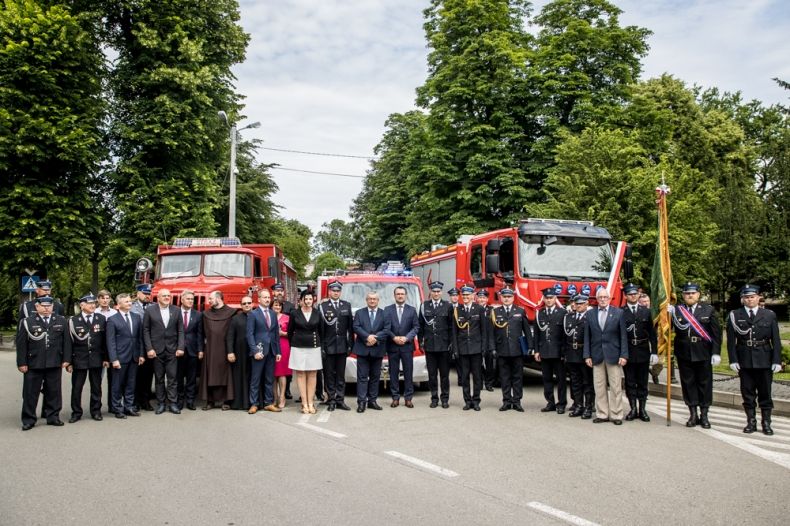 Wicemarszałek Łukasz Smółka stoi z pozostałymi uczestnikami wydarzenia. Z tyłu widoczne wozy strażackie i zieleń.