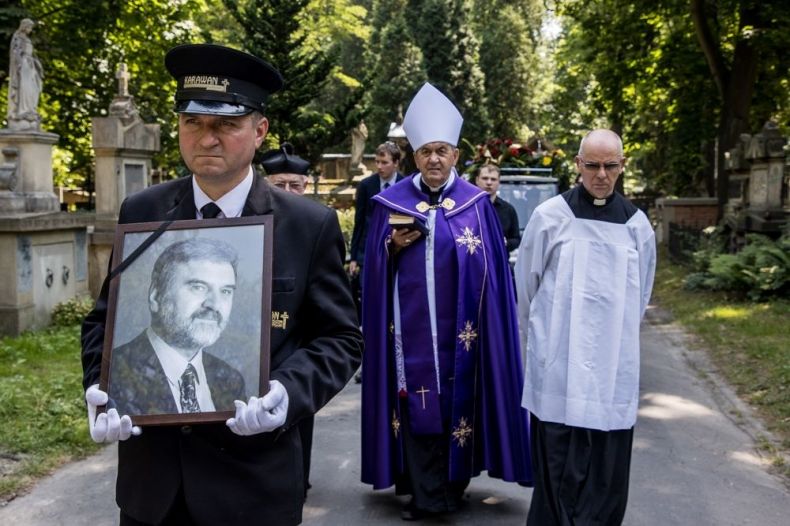 Kondukt pogrzebowy idzie alejką Cmentarza Rakowickiego. Na pierwszym planie pracownik zakładu niesie zdjęcie zmarłego, z tyłu idzie infułat Bronisław Fidelus i ksiądz.